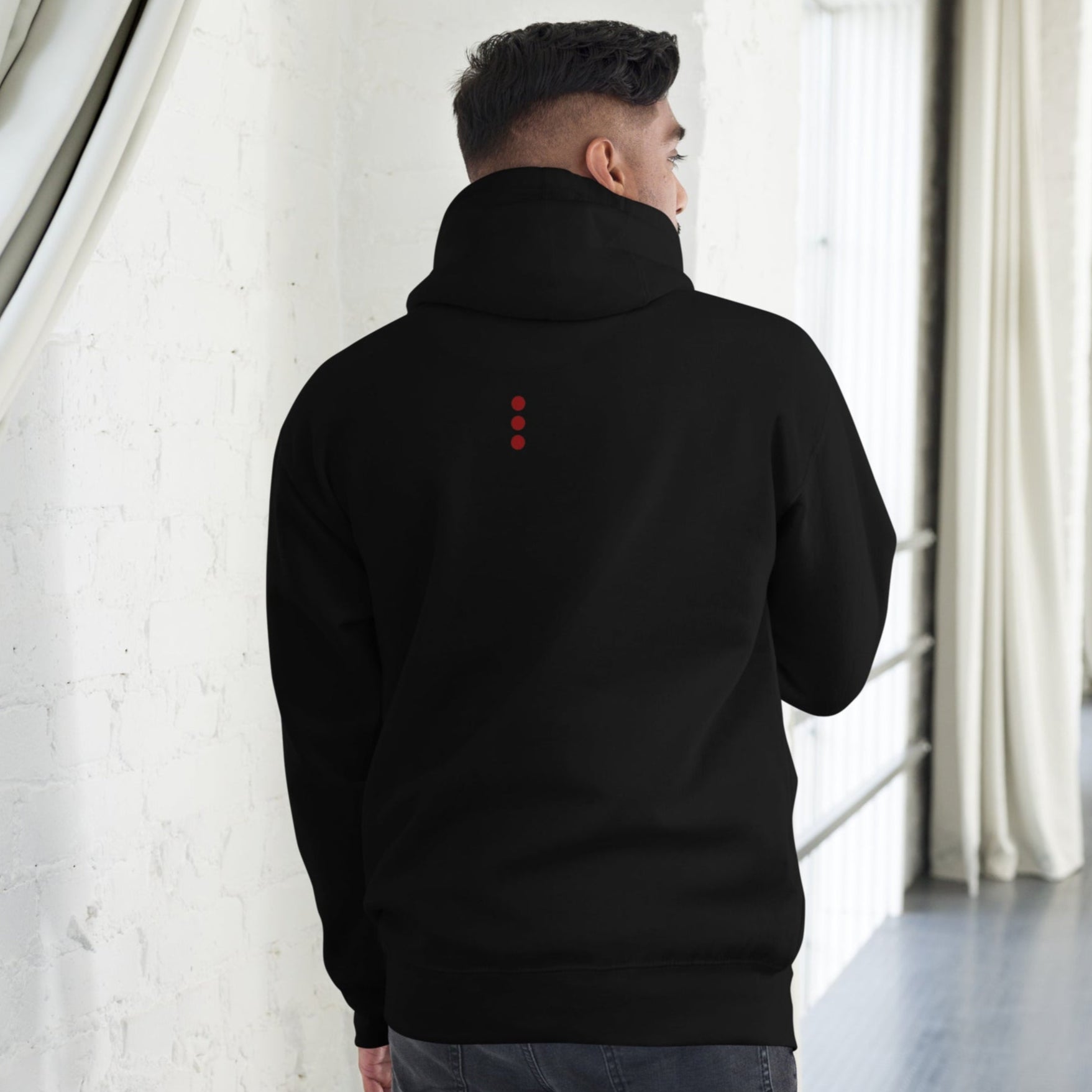 Person wearing a black hoodie with a red logo on the back, standing in a room with white curtains.