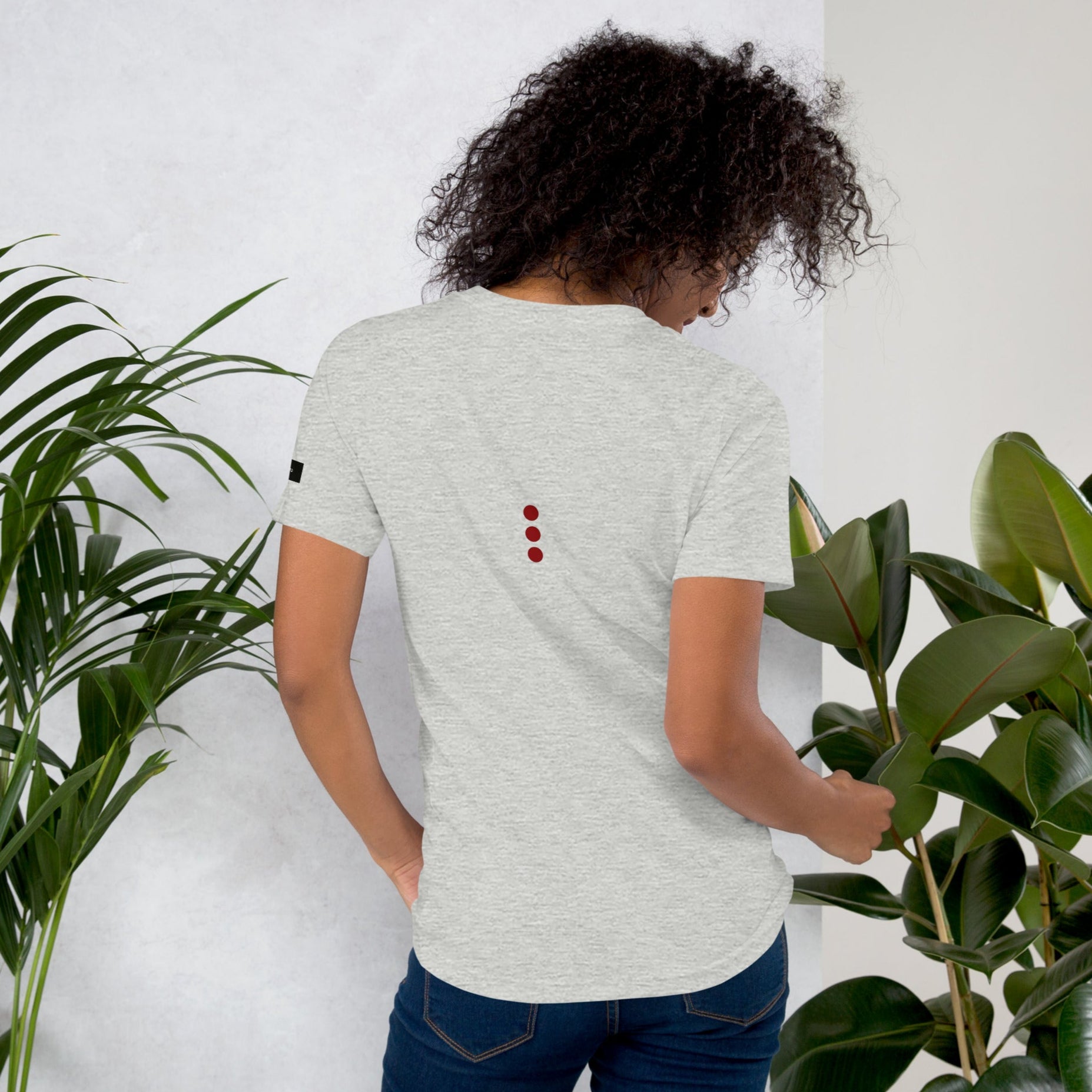 Person wearing a light gray t-shirt with red accents, standing between two plants against a white wall.
